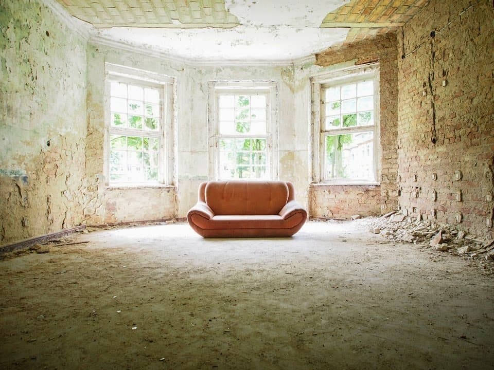 An orange sofa alone in a peeling, sunlit room