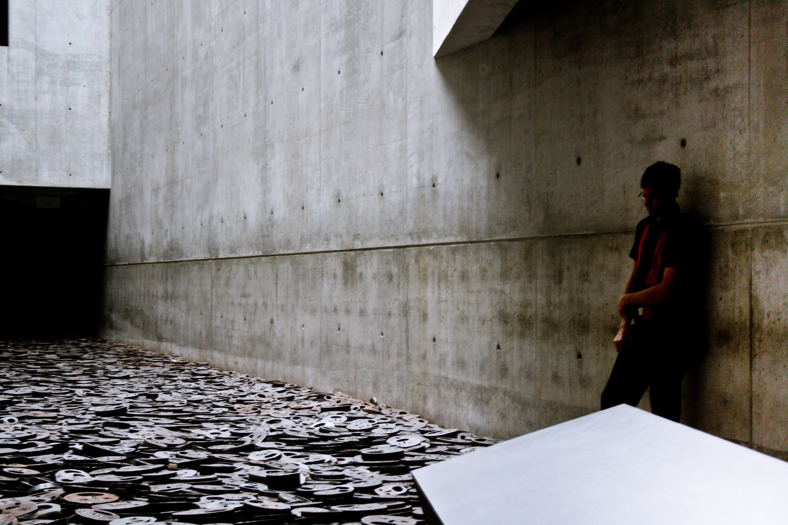 A tall concrete void with a floor of iron faces, a figure against the wall